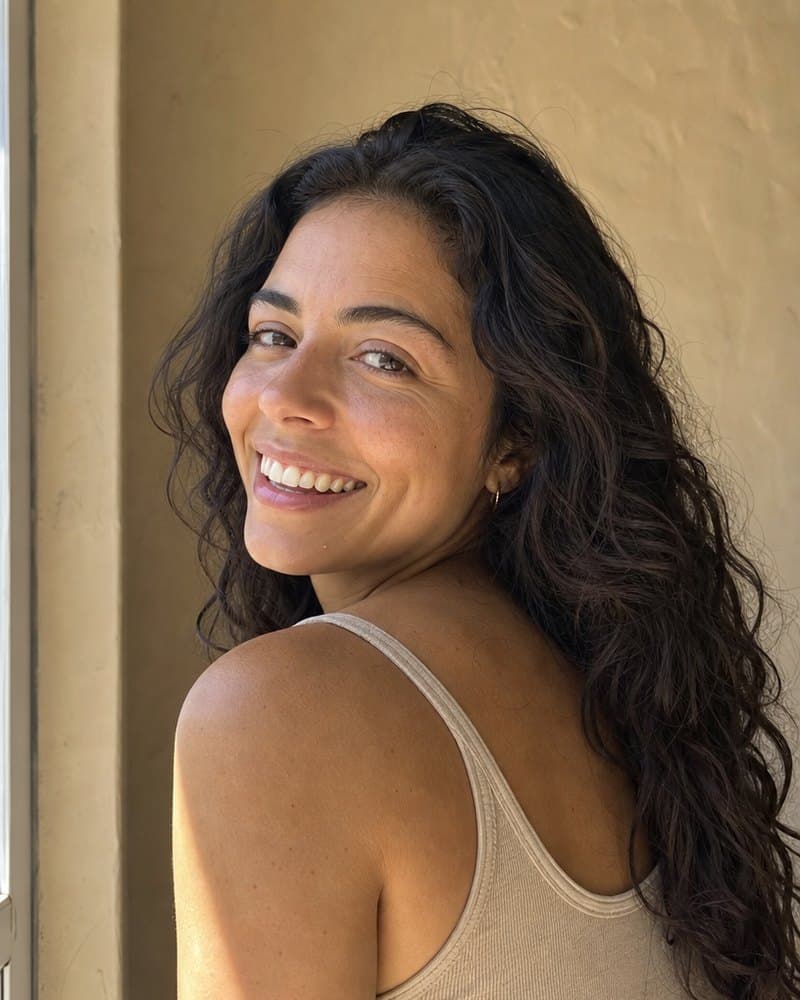 Young woman with long dark curls laughing over her shoulder in a beige tank top