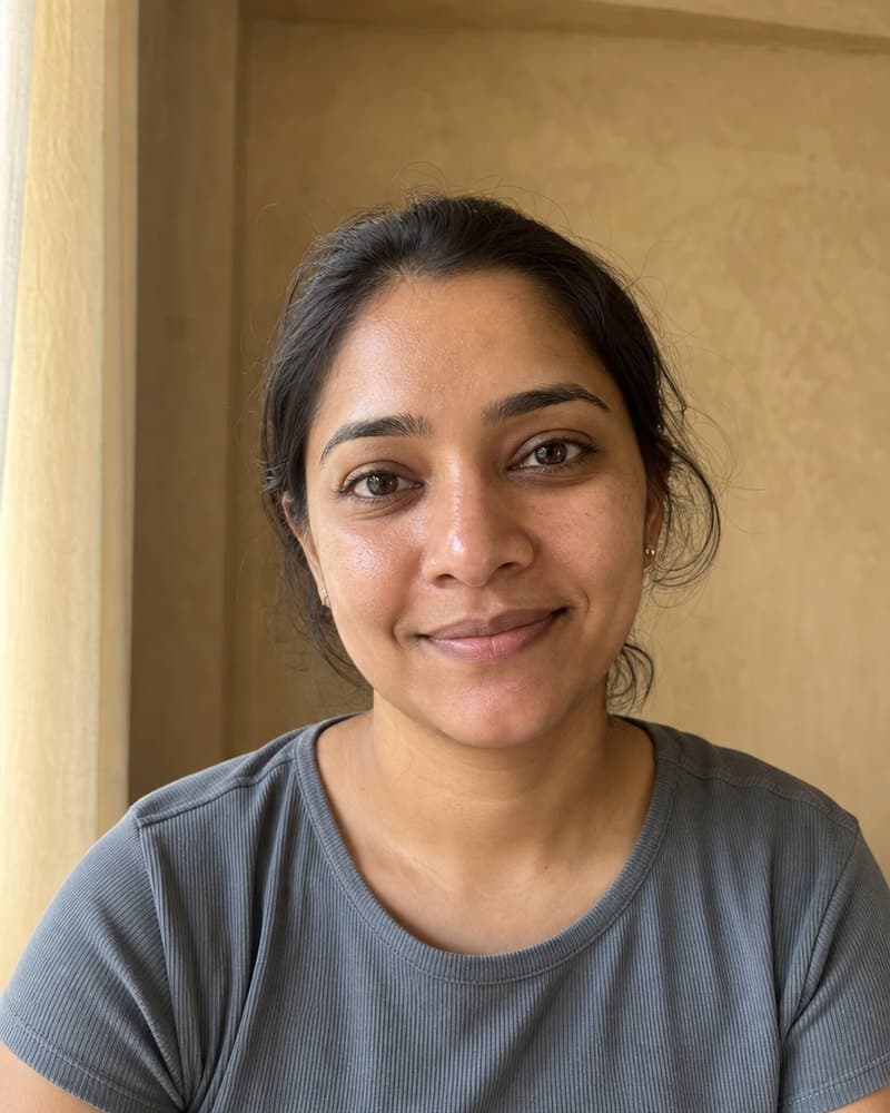 Woman in her thirties with her hair tied back and a soft smile, in a grey-blue T-shirt by a window